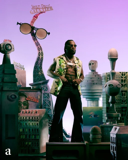 Adult male poses in a green vest, black pants, and jewelry amid surreal city backdrop with Eiffel Tower and Jean Paul Gaultier signage.