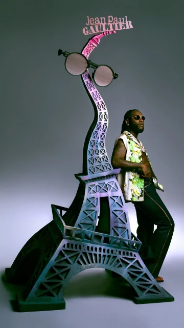Man in patterned shirt poses next to an Eiffel Tower statue adorned with sunglasses and a Jean Paul Gaultier sign against grey backdrop.