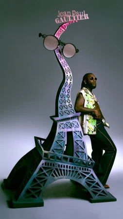 Man in patterned shirt poses next to an Eiffel Tower statue adorned with sunglasses and a Jean Paul Gaultier sign against grey backdrop.