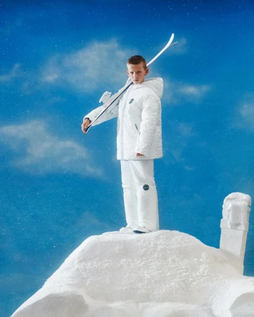 Young boy in white snowsuit and skis posing on faux snow hill against blue sky with clouds and snow dust effect, winter fashion
