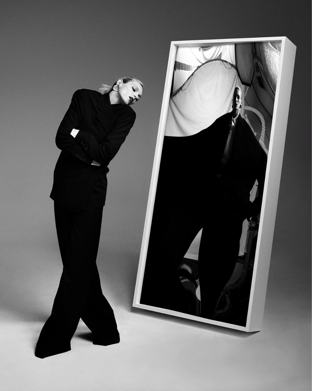 Black and white fashion shot of a woman in a dark suit leaning next to a framed image of a man in a similar suit on a grey background.