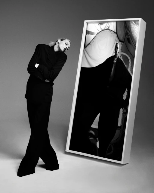Black and white fashion shot of a woman in a dark suit leaning next to a framed image of a man in a similar suit on a grey background.