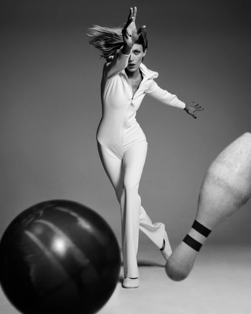 A woman in a white jumpsuit throws a bowling ball at a pin in a black and white studio shot with a neutral gray background.