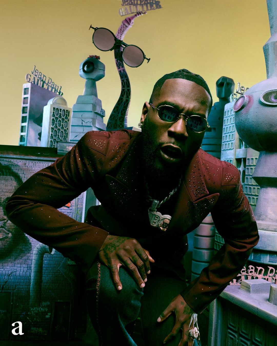 Portrait of a stylish adult male crouched, wearing sunglasses, a maroon jacket, and jewelry, with building and structure illustrations in the background.