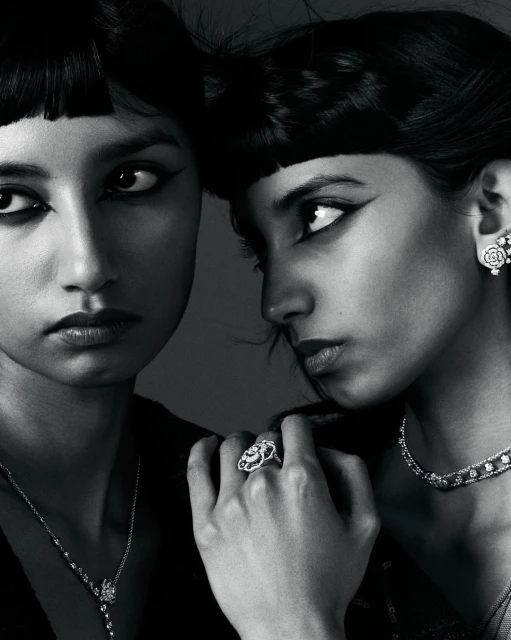 Close-up studio shot of two adult women wearing elegant jewelry and black clothing, striking dark eye makeup, and sleek dark bangs.