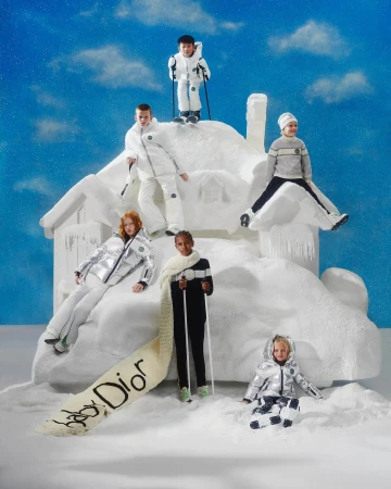 A group of six children dressed in winter ski clothing posing on a snow-covered set with a toy snow-covered house against a blue sky.