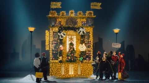 Festive scene with people in front of a Christmas-themed building facade illuminated with fairy lights, featuring holiday decorations and shopping bags.