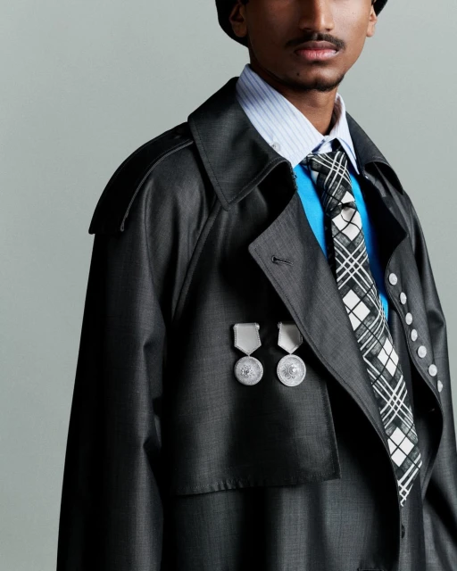 Stylish individual in a dark trench coat with military-style badges, accessorized with a patterned tie and matching hat. Fashion editorial.
