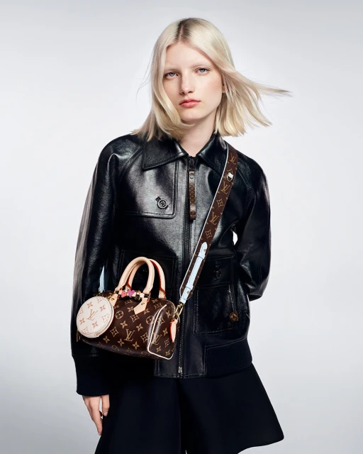 Fashion shot of a fair-skinned model with blonde hair wearing a black leather jacket, holding a brown Louis Vuitton purse, in studio.