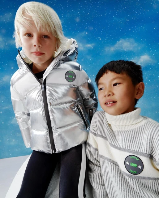 Two children wearing winter clothing pose against a sky-blue backdrop with clouds and light snowfall. The children are wearing Dior-branded clothing.