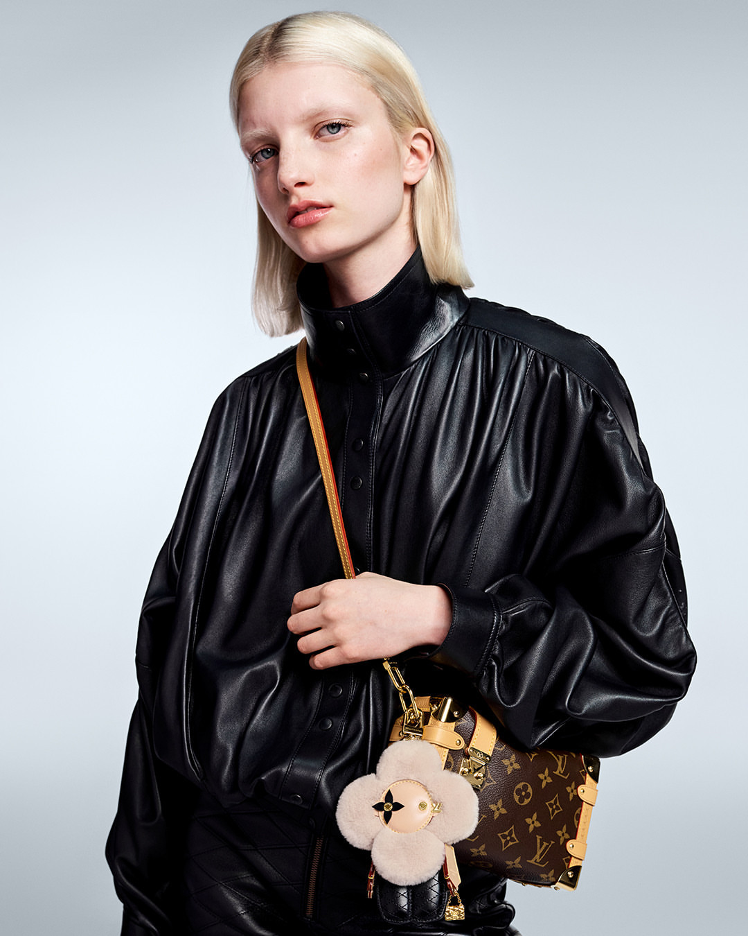 Fashion shot of a blonde woman in a black leather jacket holding a Louis Vuitton handbag with a furry flower charm in a studio.