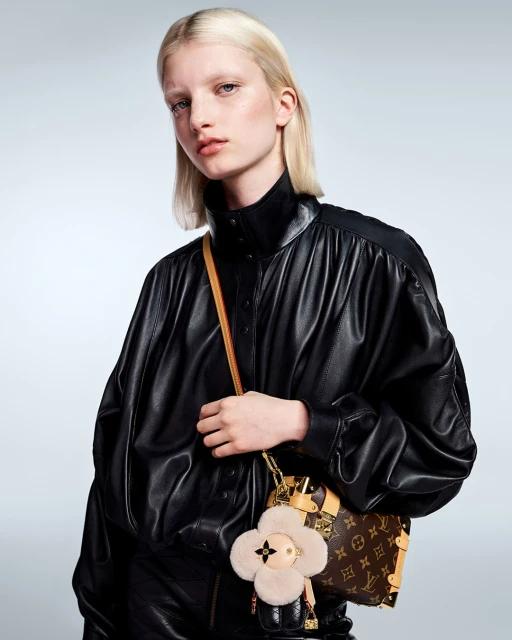 Fashion shot of a blonde woman in a black leather jacket holding a Louis Vuitton handbag with a furry flower charm in a studio.