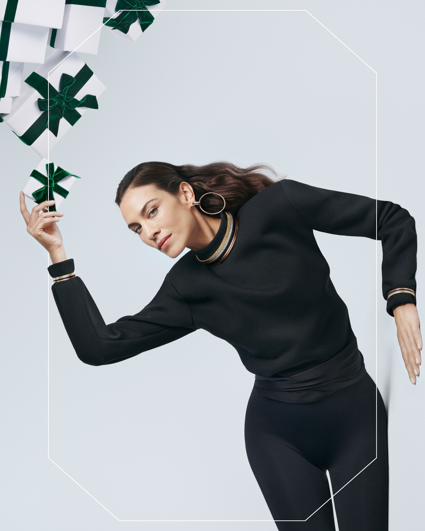 Brunette woman in black sweater and leggings poses near white gift boxes with green ribbons in a fashion-style indoor setting.