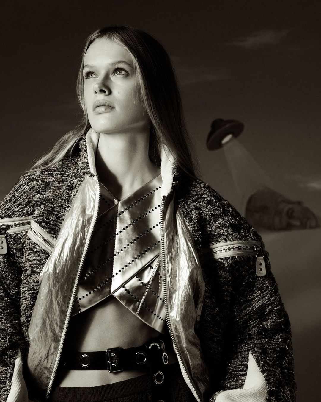 A woman with long hair poses in a jacket and top, looking upward, with an object resembling a UFO in the sepia-toned sky background.