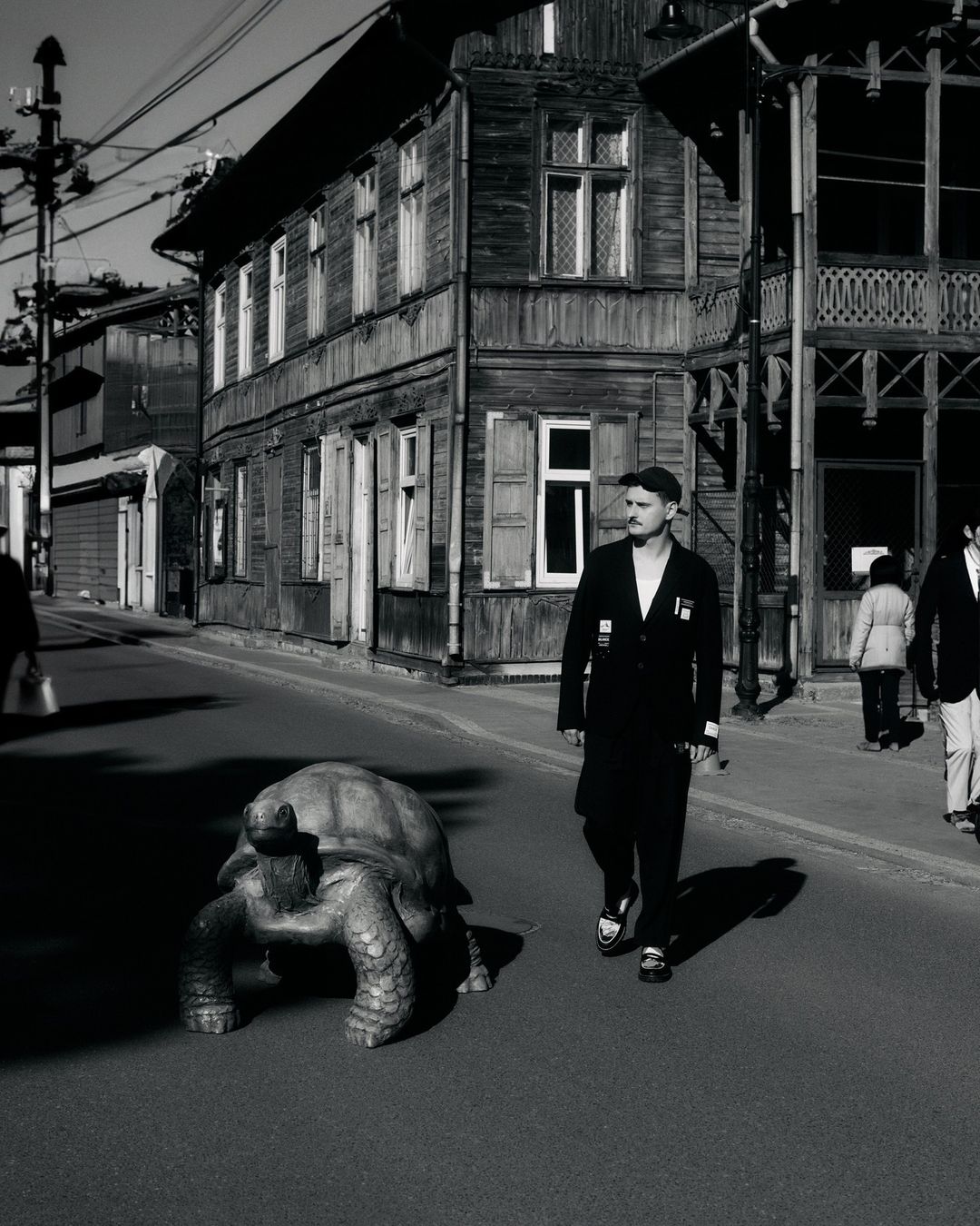Monochrome photo of a man walking a giant tortoise on a street lined with wooden buildings, creating a vintage and surreal scene.