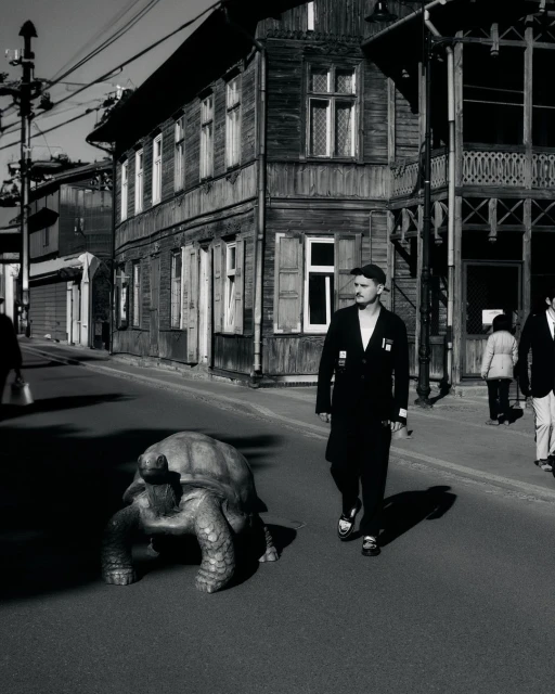 Monochrome photo of a man walking a giant tortoise on a street lined with wooden buildings, creating a vintage and surreal scene.