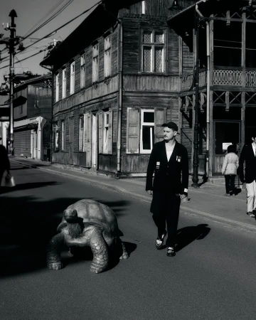 Monochrome photo of a man walking a giant tortoise on a street lined with wooden buildings, creating a vintage and surreal scene.