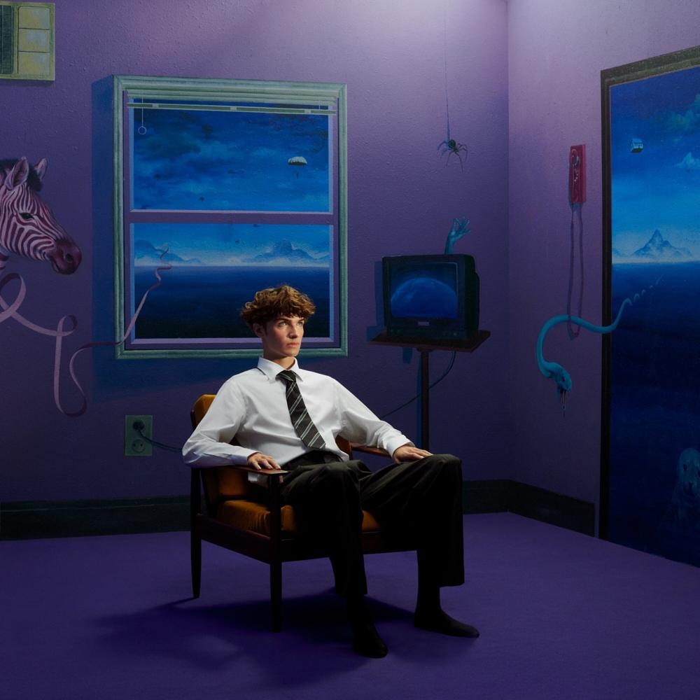 Young man in shirt and tie sitting in chair in surreal violet room with zebra head, paintings, retro TV, and hanging spider.