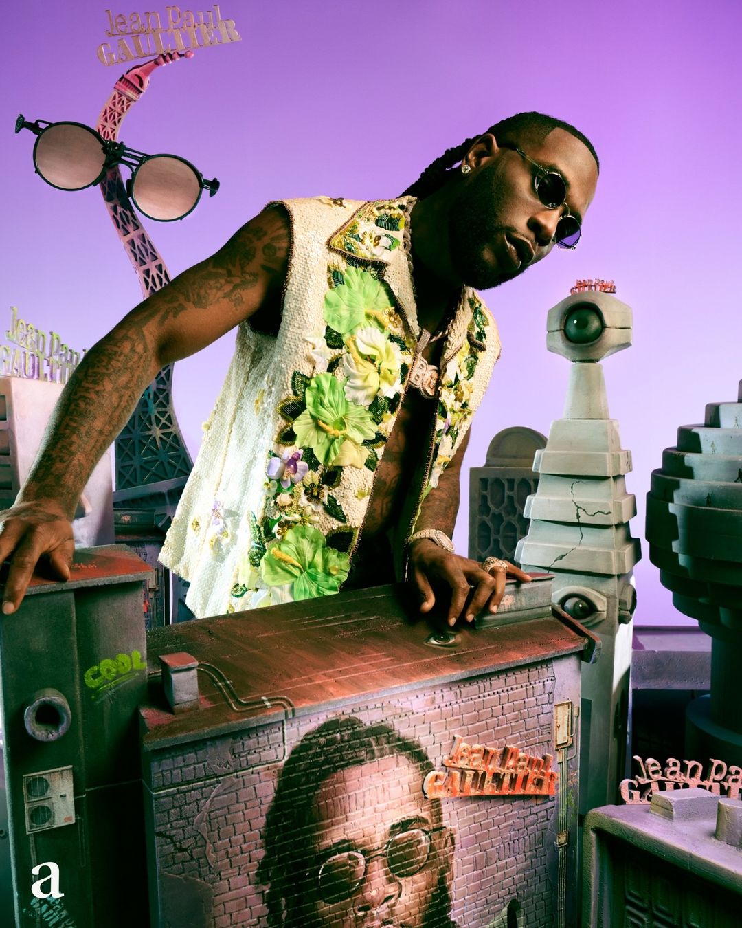 Burna Boy in a sleeveless floral embroidered top posing among surreal miniature architectural props against a purple backdrop.