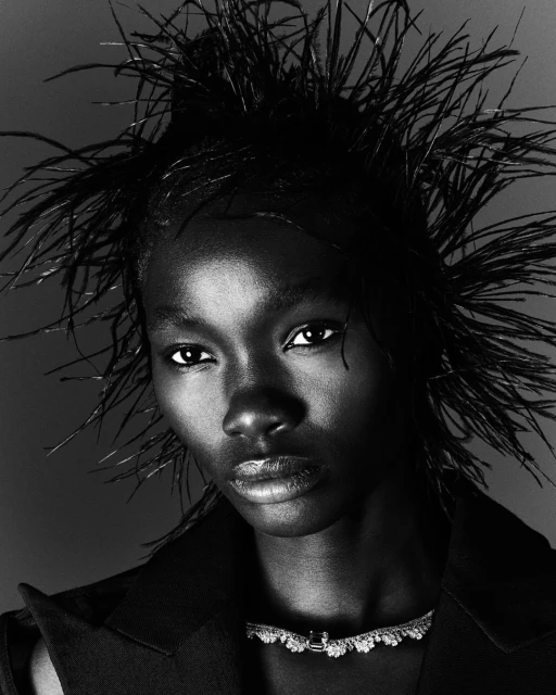 Close-up black-and-white portrait of an adult female model with dark skin and feather headdress, wearing jewelry and a jacket.