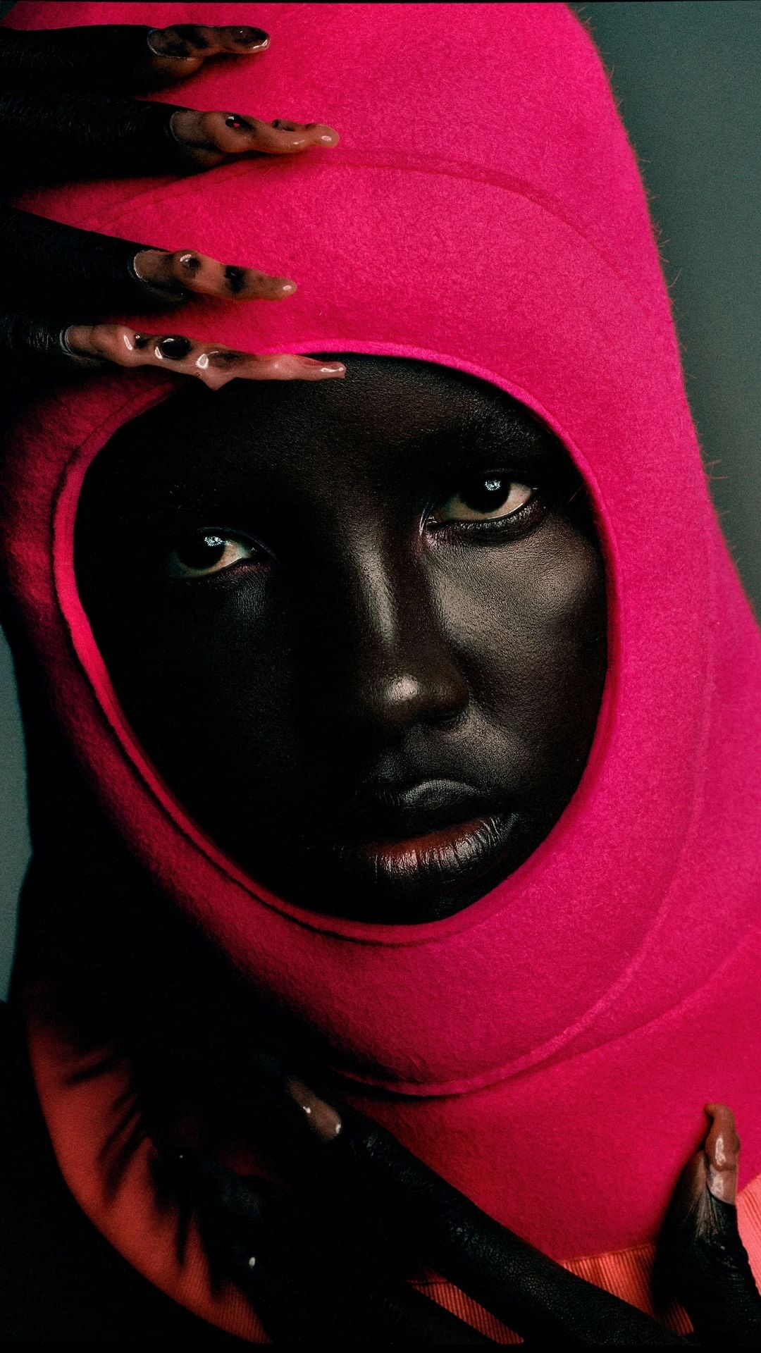 Close-up portrait of an African woman with painted nails and a pink hood, showcasing fashion and beauty against a blue background.