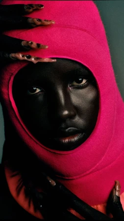 Close-up portrait of an African woman with painted nails and a pink hood, showcasing fashion and beauty against a blue background.