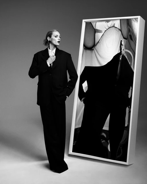 Monochrome fashion portrait of an adult female in an oversized suit standing beside a framed image on a grey gradient background.