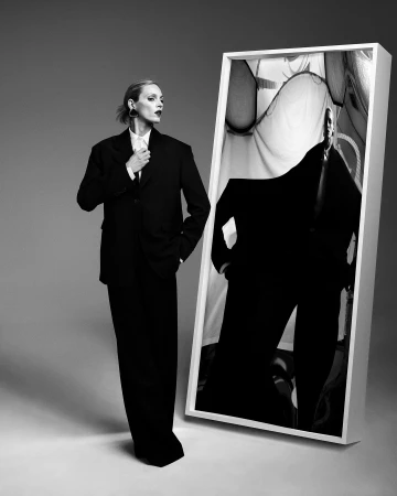 Monochrome fashion portrait of an adult female in an oversized suit standing beside a framed image on a grey gradient background.