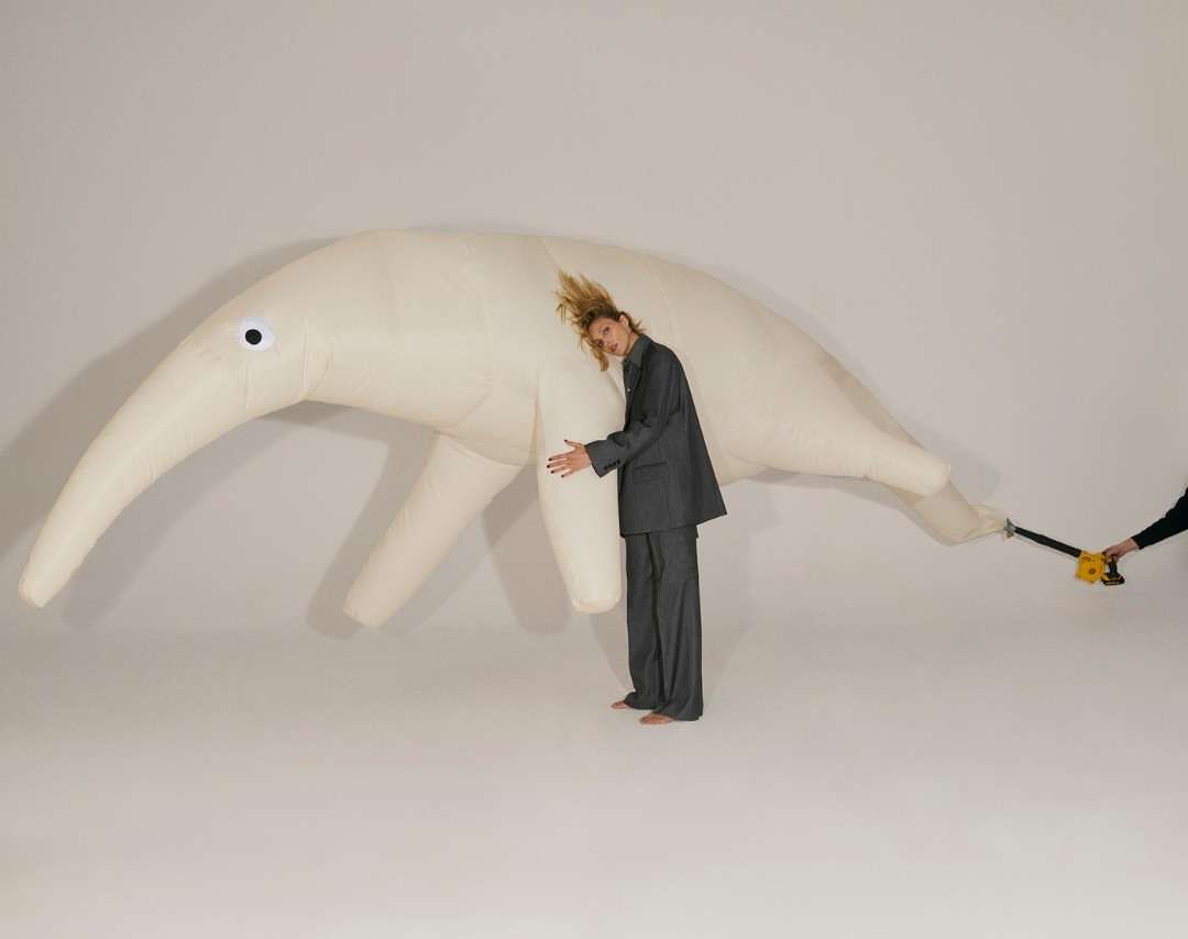 Adult woman in a grey suit hugs a giant inflatable anteater in a white studio. Someone off-camera holds a leaf blower.