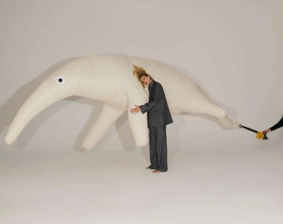 Adult woman in a grey suit hugs a giant inflatable anteater in a white studio. Someone off-camera holds a leaf blower.