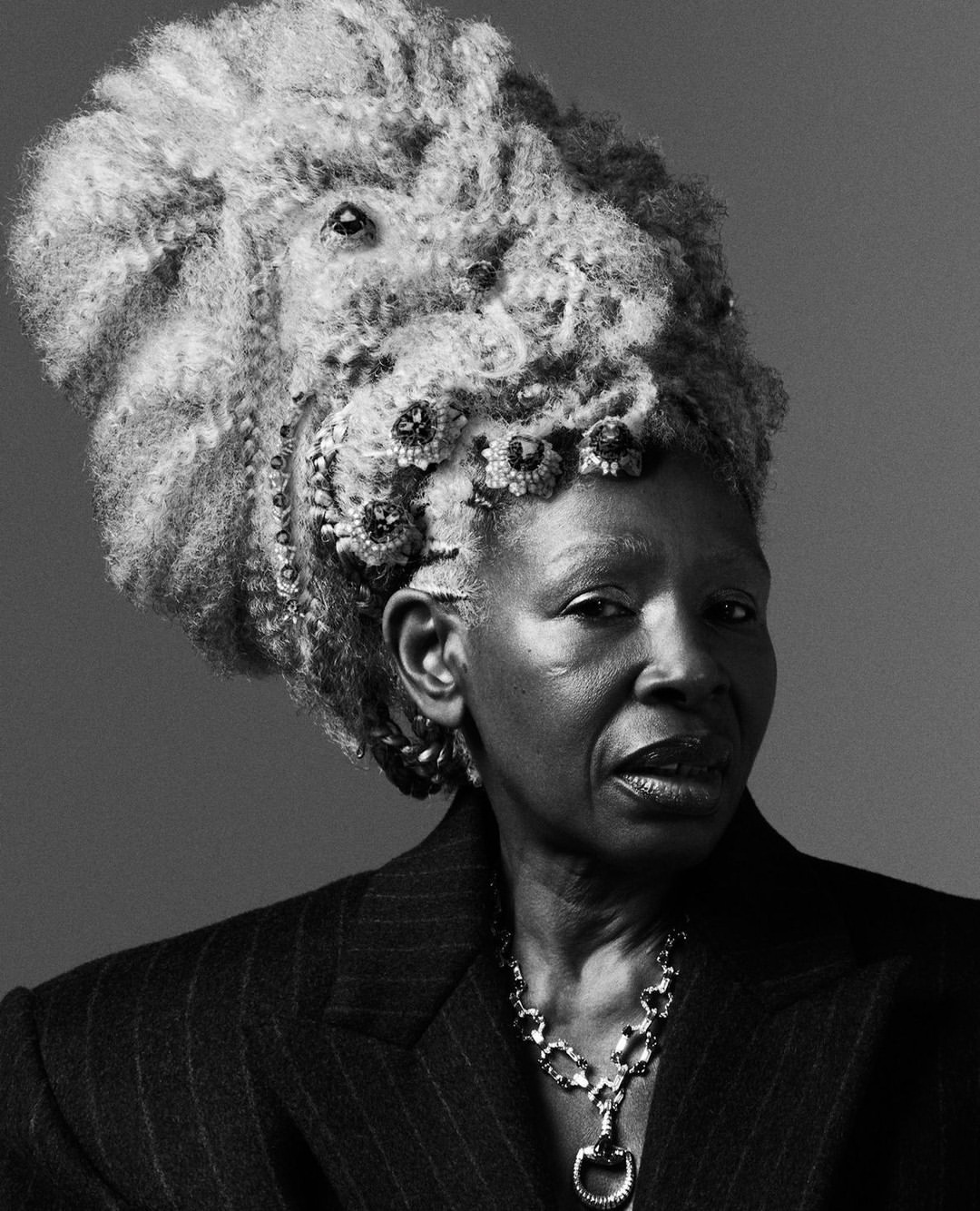 Headshot of mature Black woman with lion-like hair creation, pinstripe jacket, and statement necklace against a neutral background.