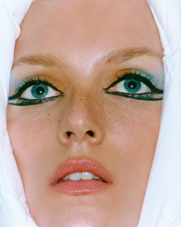 Close-up portrait of a woman with blue eyes, dramatic black winged eyeliner, blue eyeshadow, and freckles, framed by white fabric.