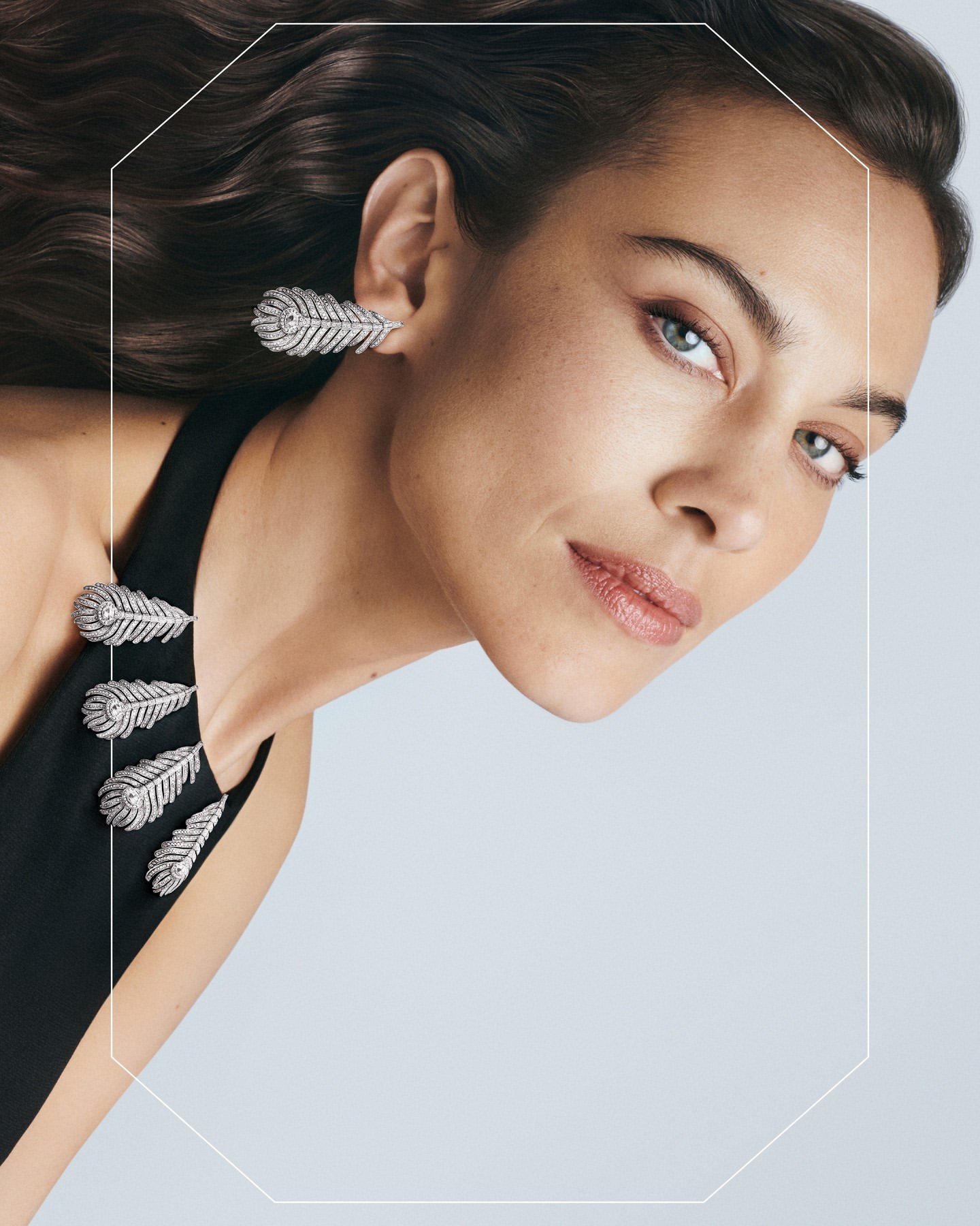 Portrait of adult woman wearing black halter dress and feather-shaped jewelry earrings and necklace, against a pale background.