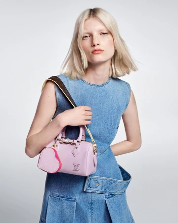 Blonde female model wearing a blue denim dress holding a light pink handbag with a heart coin purse.