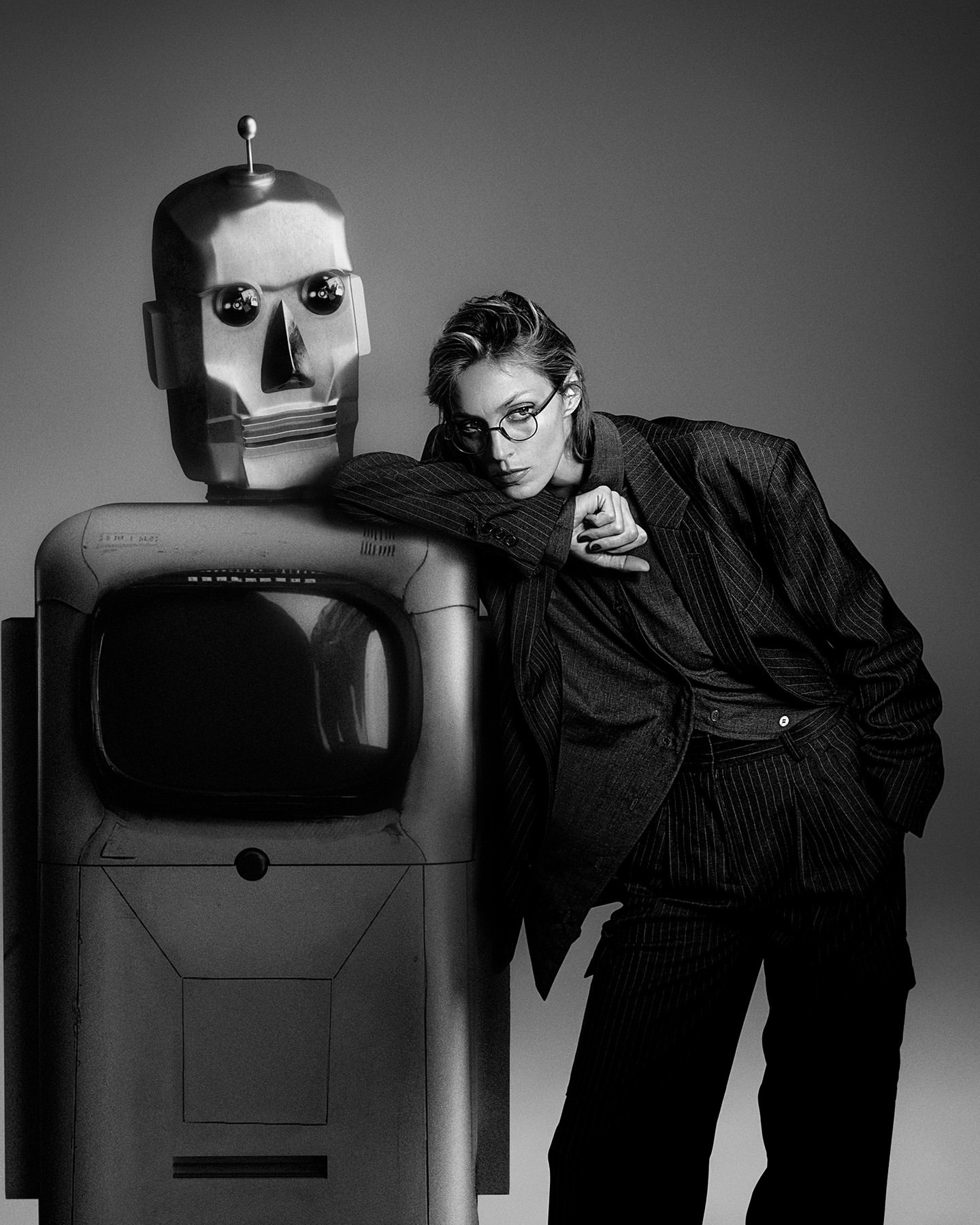 A black-and-white fashion shot of a woman in a pinstripe suit leaning against a robot with a vintage television screen for a body.