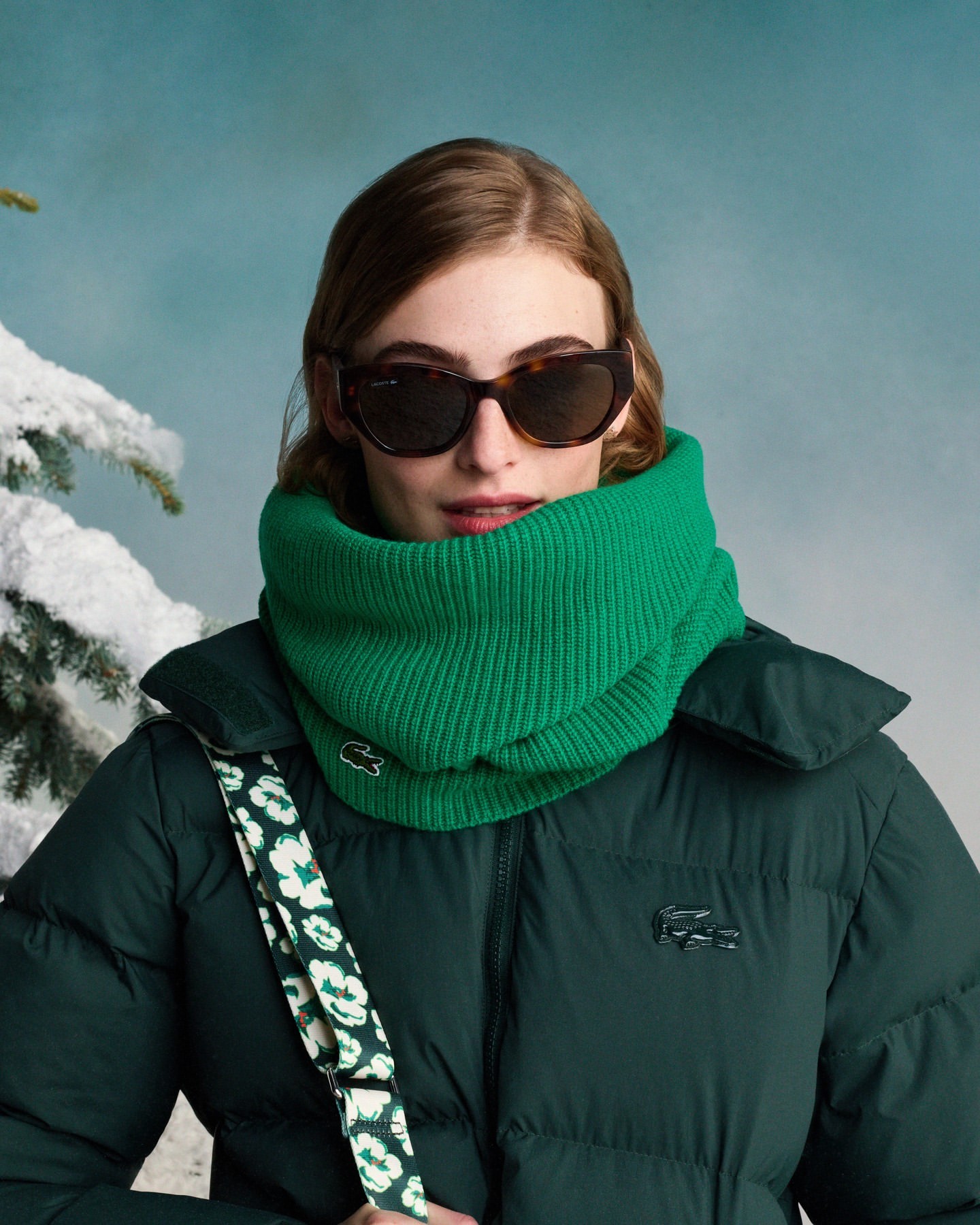 A stylish young woman in sunglasses and a Lacoste puffy coat and scarf poses against a wintery blue sky backdrop with snow-covered tree.