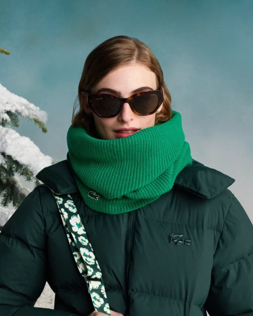 A stylish young woman in sunglasses and a Lacoste puffy coat and scarf poses against a wintery blue sky backdrop with snow-covered tree.