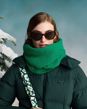 A stylish young woman in sunglasses and a Lacoste puffy coat and scarf poses against a wintery blue sky backdrop with snow-covered tree.