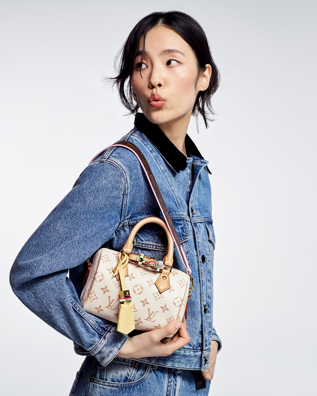 Asian woman in denim jacket and jeans poses in a white studio, holding a Louis Vuitton handbag with monogram.