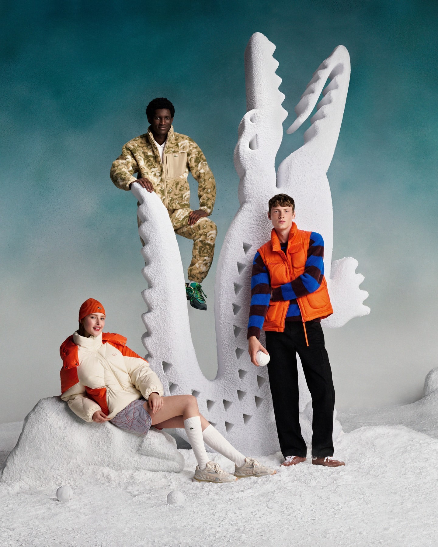 Three fashion models pose in a stylized outdoor winter scene with a large decorative crocodile, wearing outerwear and holding snowballs.