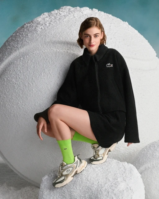 A woman in a black jacket and skirt poses next to a textured white ball wearing green socks and fashion sneakers in a studio setting.