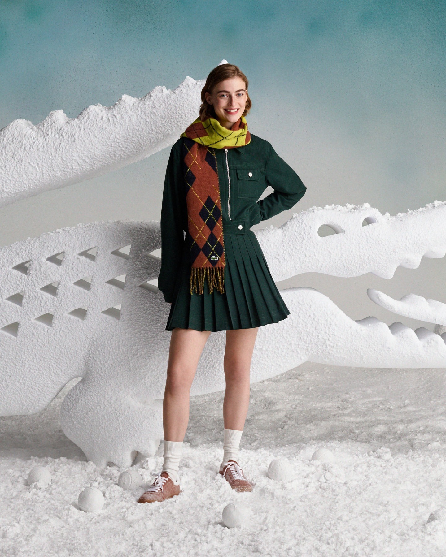 Smiling woman poses in a green jacket and skirt with a patterned scarf, standing in a wintery scene with a white crocodile prop.