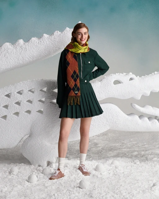 Smiling woman poses in a green jacket and skirt with a patterned scarf, standing in a wintery scene with a white crocodile prop.