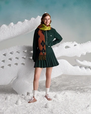 Smiling woman poses in a green jacket and skirt with a patterned scarf, standing in a wintery scene with a white crocodile prop.