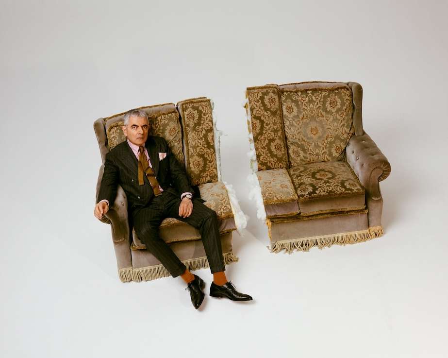 Elegant man in a suit sits in half a deconstructed armchair against a white backdrop, blending classic style with a modern deconstructed twist.