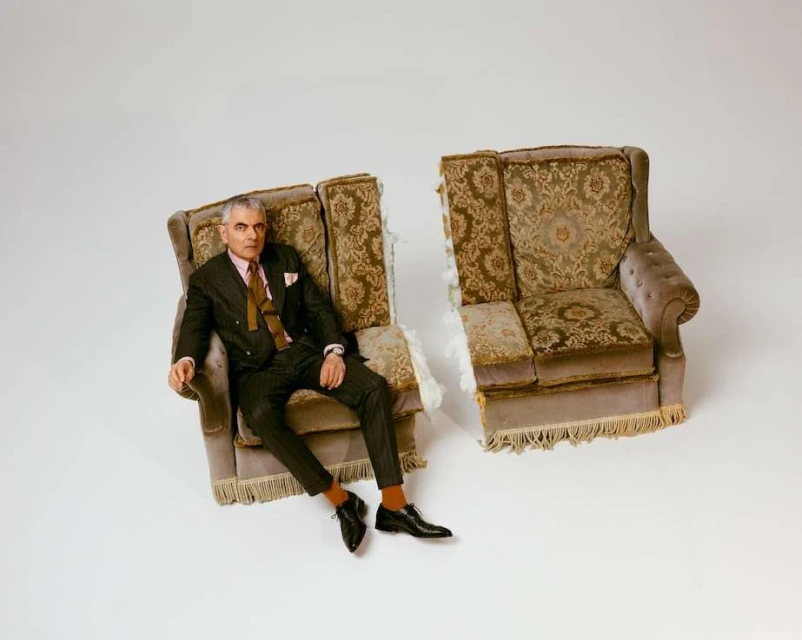 Elegant man in a suit sits in half a deconstructed armchair against a white backdrop, blending classic style with a modern deconstructed twist.