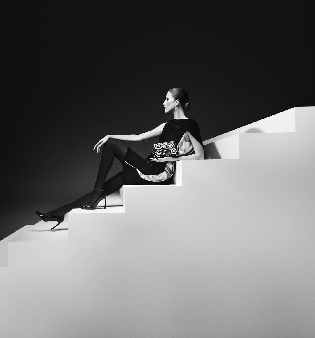 Monochrome fashion shot of a woman seated on white stairs holding a floral handbag against a dark background.