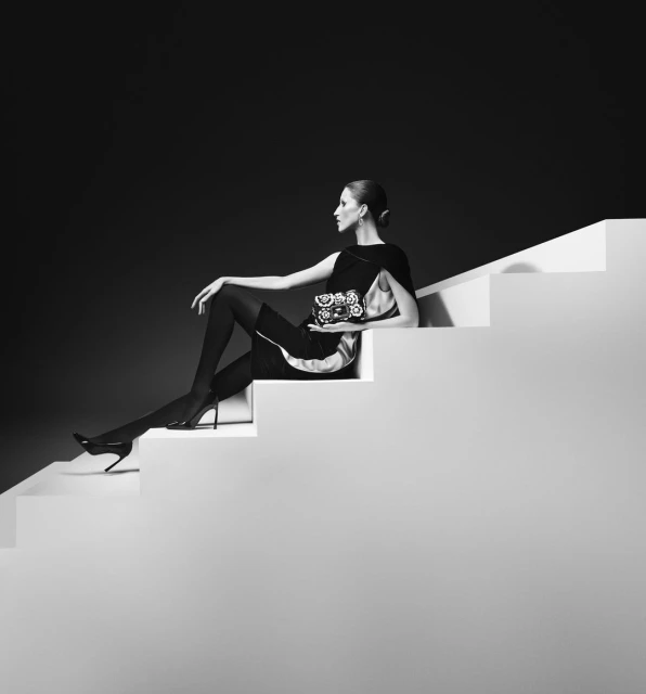 Monochrome fashion shot of a woman seated on white stairs holding a floral handbag against a dark background.