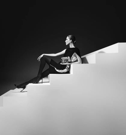 Monochrome fashion shot of a woman seated on white stairs holding a floral handbag against a dark background.