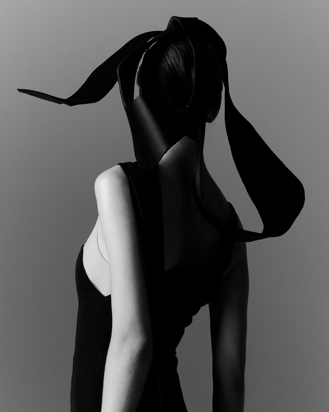 Monochrome studio portrait of a woman with dark hair, wearing a dark sleeveless dress and sculptural headpiece with dramatic lighting.